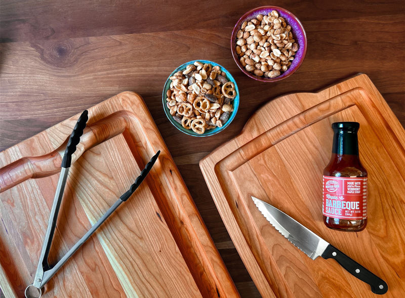 An overhead flat-lay of wooden cutting boards on a dark wood table, with a knife, a bottle of barbecue sauce, and two bowls of snacks.
