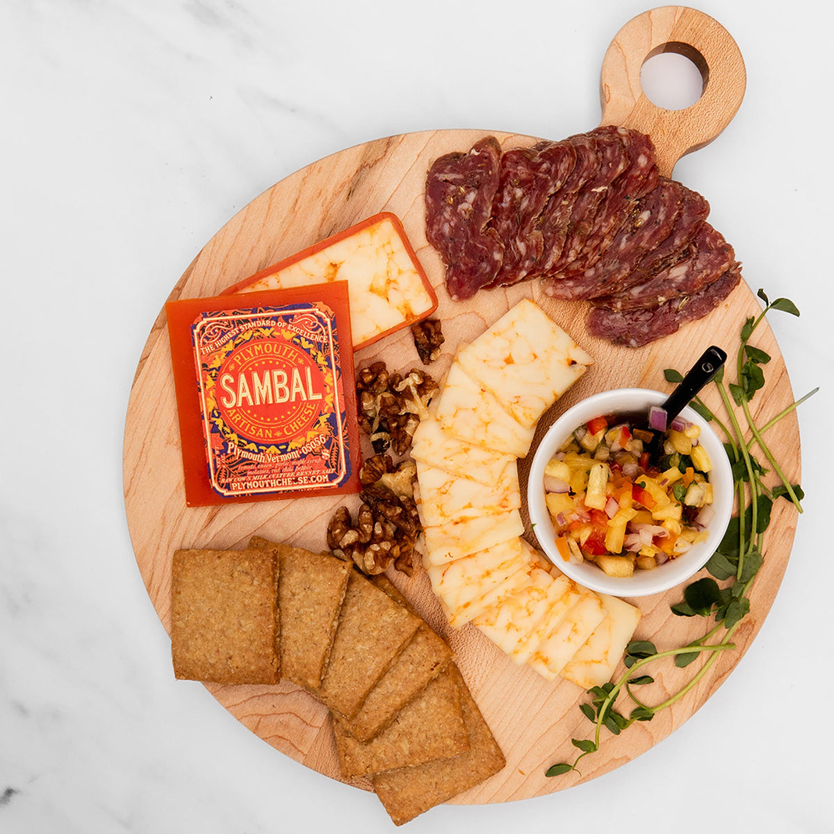 Wooden platter with cheese, salami, crackers, and a small bowl of salsa on a white background.