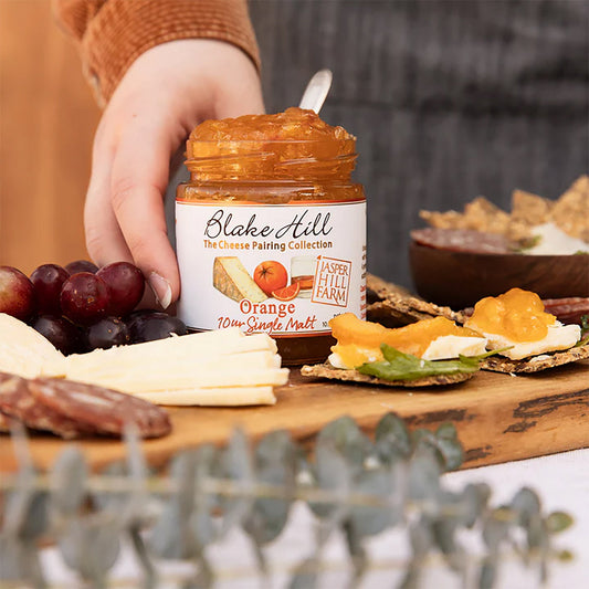 Person holding a jar of orange spread on a charcuterie board with fruits and crackers.