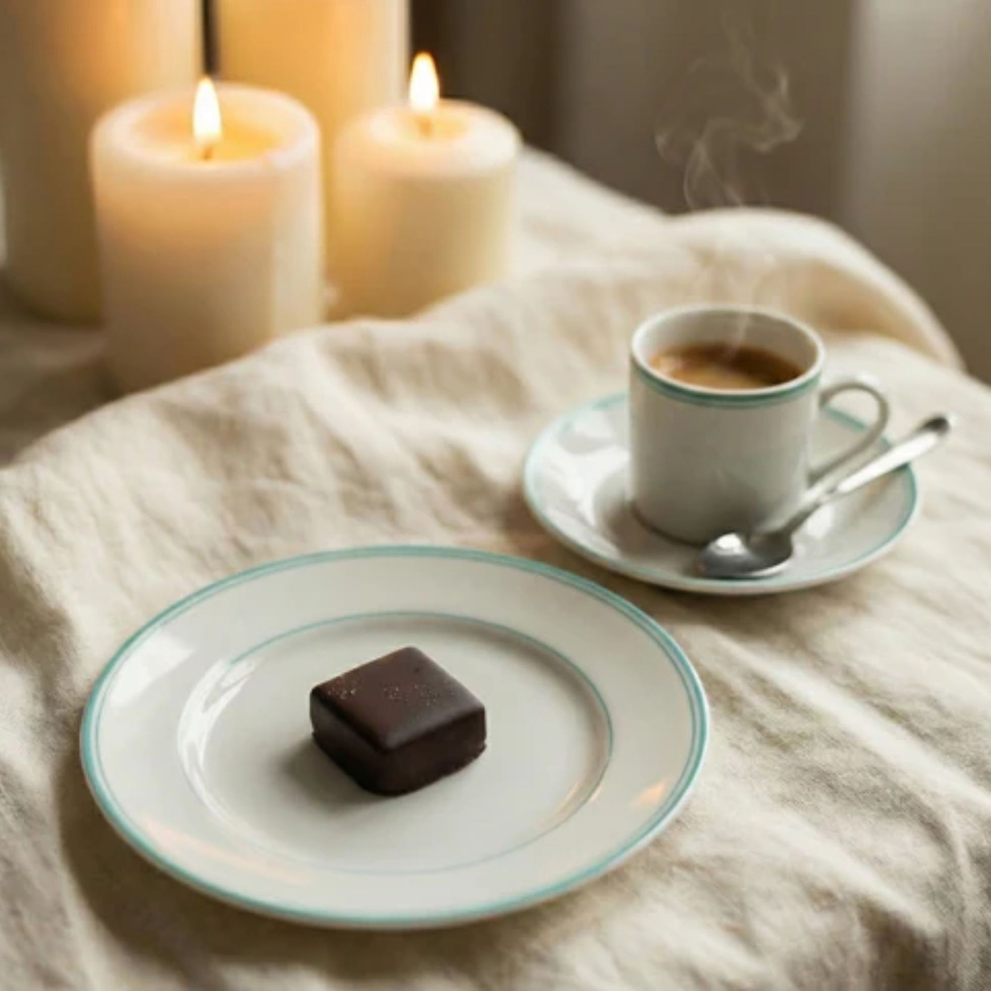 Cup of coffee with a chocolate bar on a saucer, surrounded by lit candles on a soft surface.