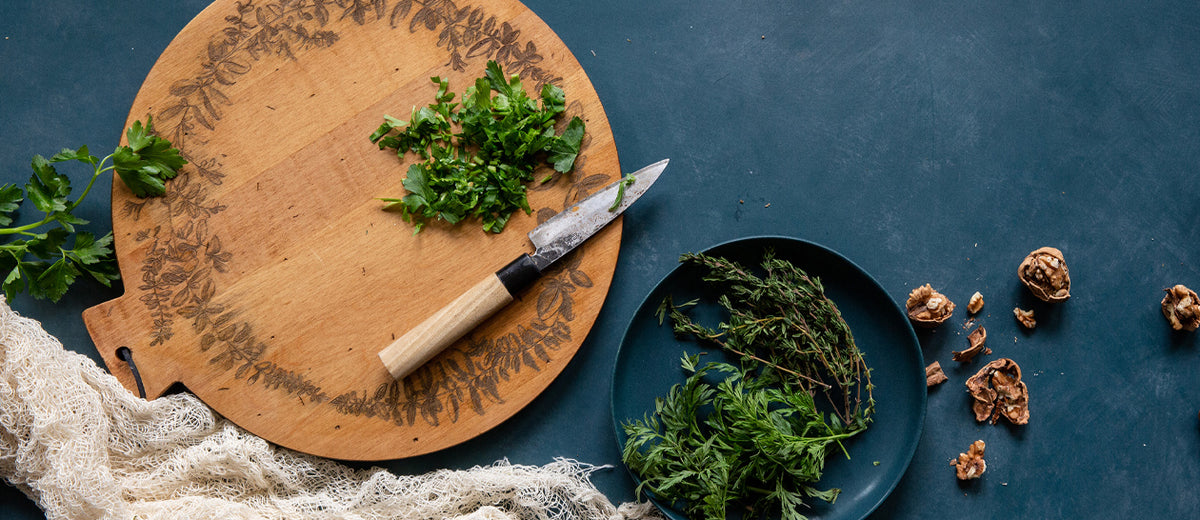 A styled flat-lay of a round wooden cutting board with engraved leaf patterns, featuring a small knife with a light wood handle and chopped green herbs, alongside a dark plate holding fresh herbs and scattered walnut shells on a dark blue surface.