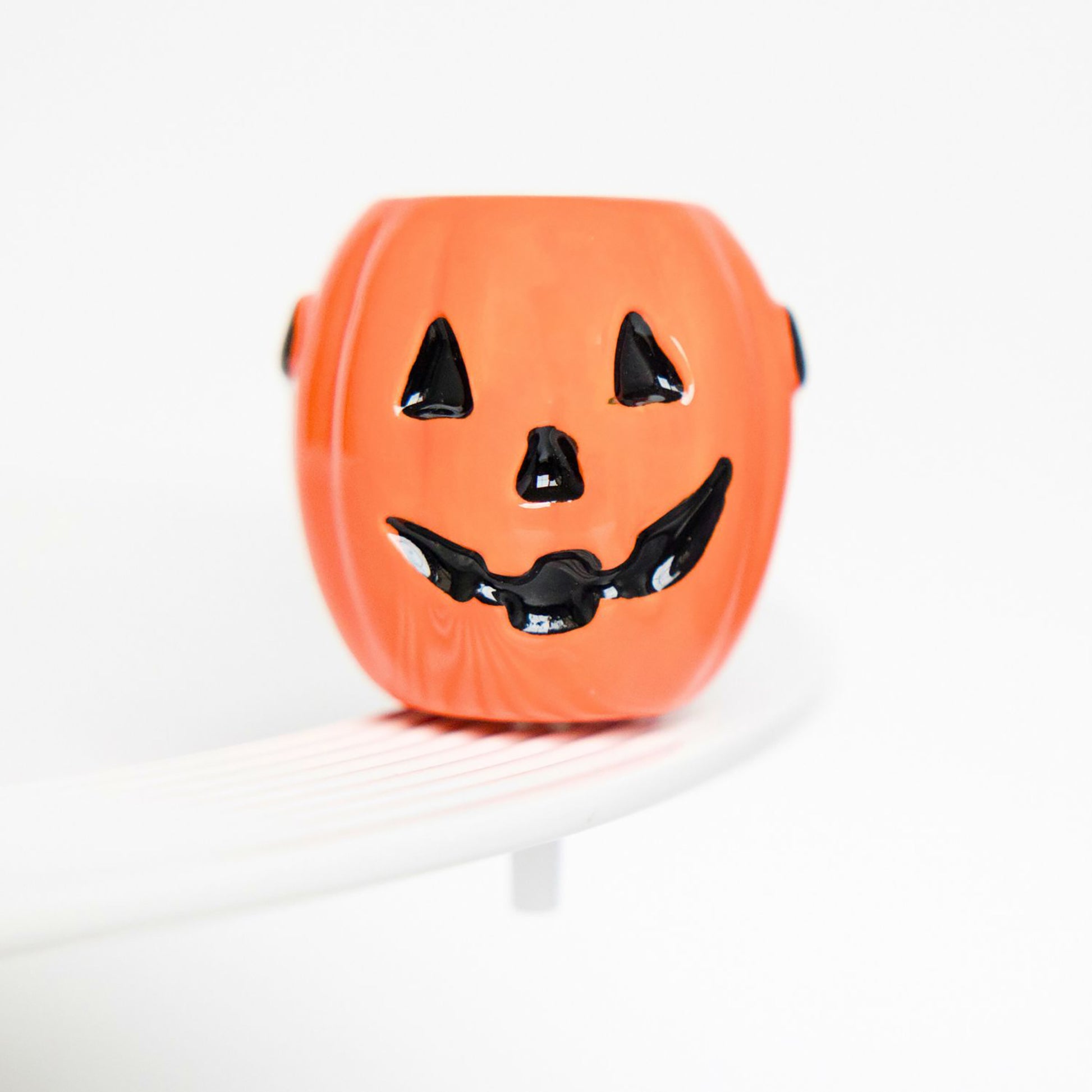 An orange jack-o'-lantern shaped container with a black painted face, sitting on a white curved surface against a clean white background.