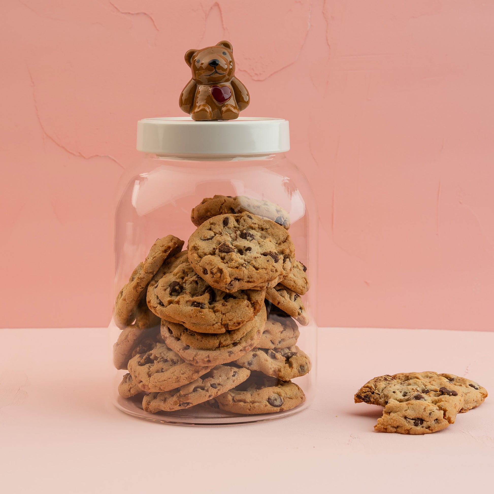 A large glass canister with a white melamine lid with a Nora Fleming teddy bear mini, filled with chocolate chip cookies. 