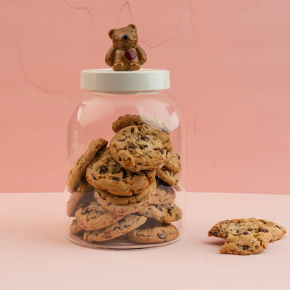 A large glass canister with a white melamine lid with a Nora Fleming teddy bear mini, filled with chocolate chip cookies. 